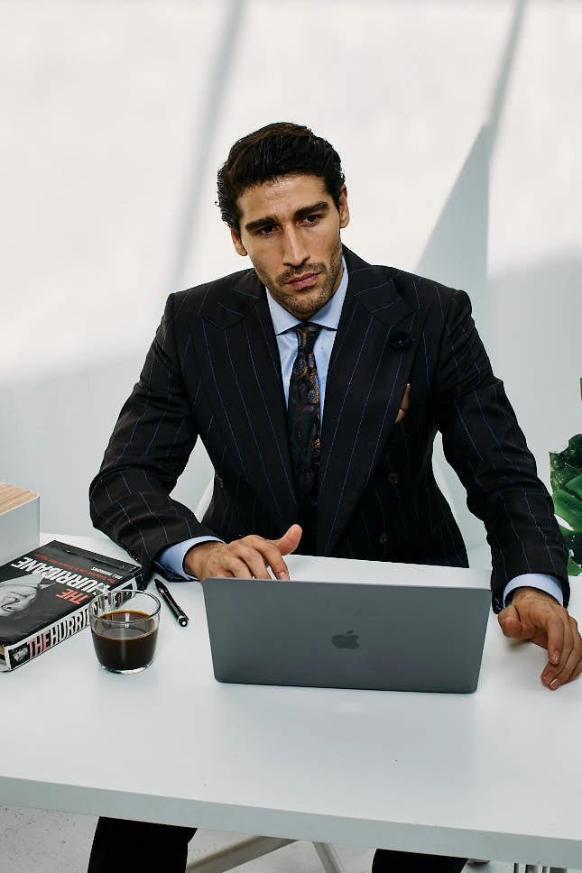 Man in a navy pinstripe double-breasted suit working on a laptop at a bright white office desk