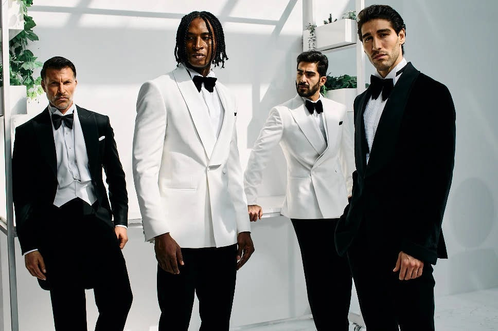 Four men in black tie, two in white dinner jackets and two in black tuxedos, in a bright studio with greenery