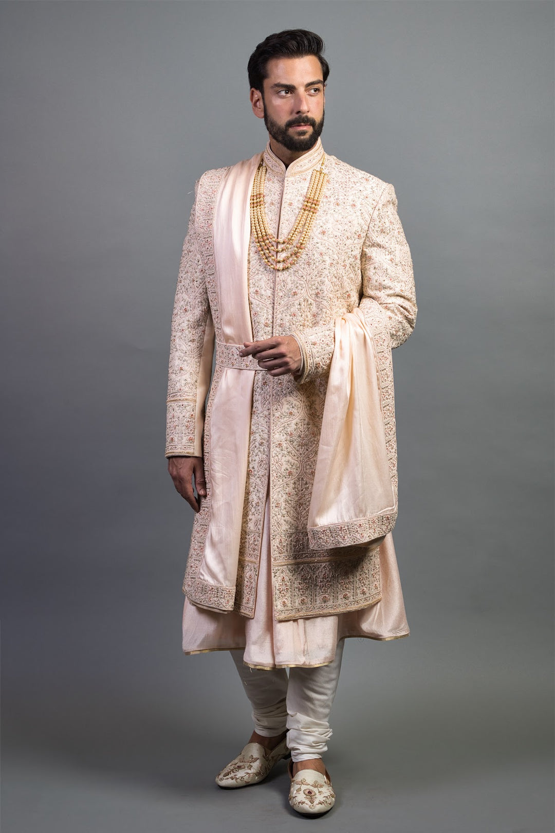 Man wearing cream and gold embroidered sherwani with matching dupatta and churidar in studio setting